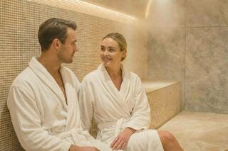 A man and woman sit in a steam room, wearing white robes, sharing a warm smile and enjoying a relaxed moment together.