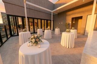 A romantic outdoor venue with elegant round tables, white tablecloths, floral centerpieces, and soft lighting, set against a sunset backdrop.