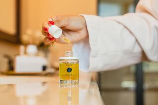 A hand with red nails holds a dropper over a yellow serum bottle, set on a sleek countertop in a softly lit space.