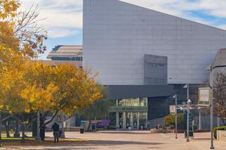 A modern science museum entrance is framed by colorful autumn trees, with clear skies and a bustling walkway leading to the entrance.