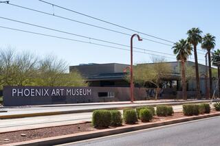 A modern building with a prominent sign reads "Phoenix Art Museum," surrounded by palm trees and desert landscaping under a clear blue sky.