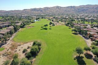 A lush green golf course stretches across the landscape, bordered by homes and mountains under a clear blue sky. Trees dot the fairway.