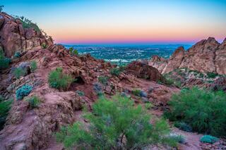 Rocky terrain with vibrant green vegetation overlooks a sprawling city at dusk, where the sky transitions from orange to blue.