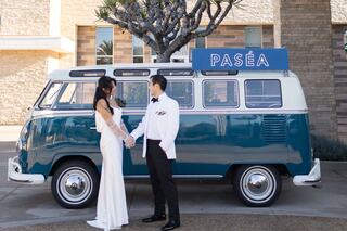 A stylish couple stands hand in hand beside a vintage blue and white van, exuding elegance and joy in a beautiful outdoor setting.